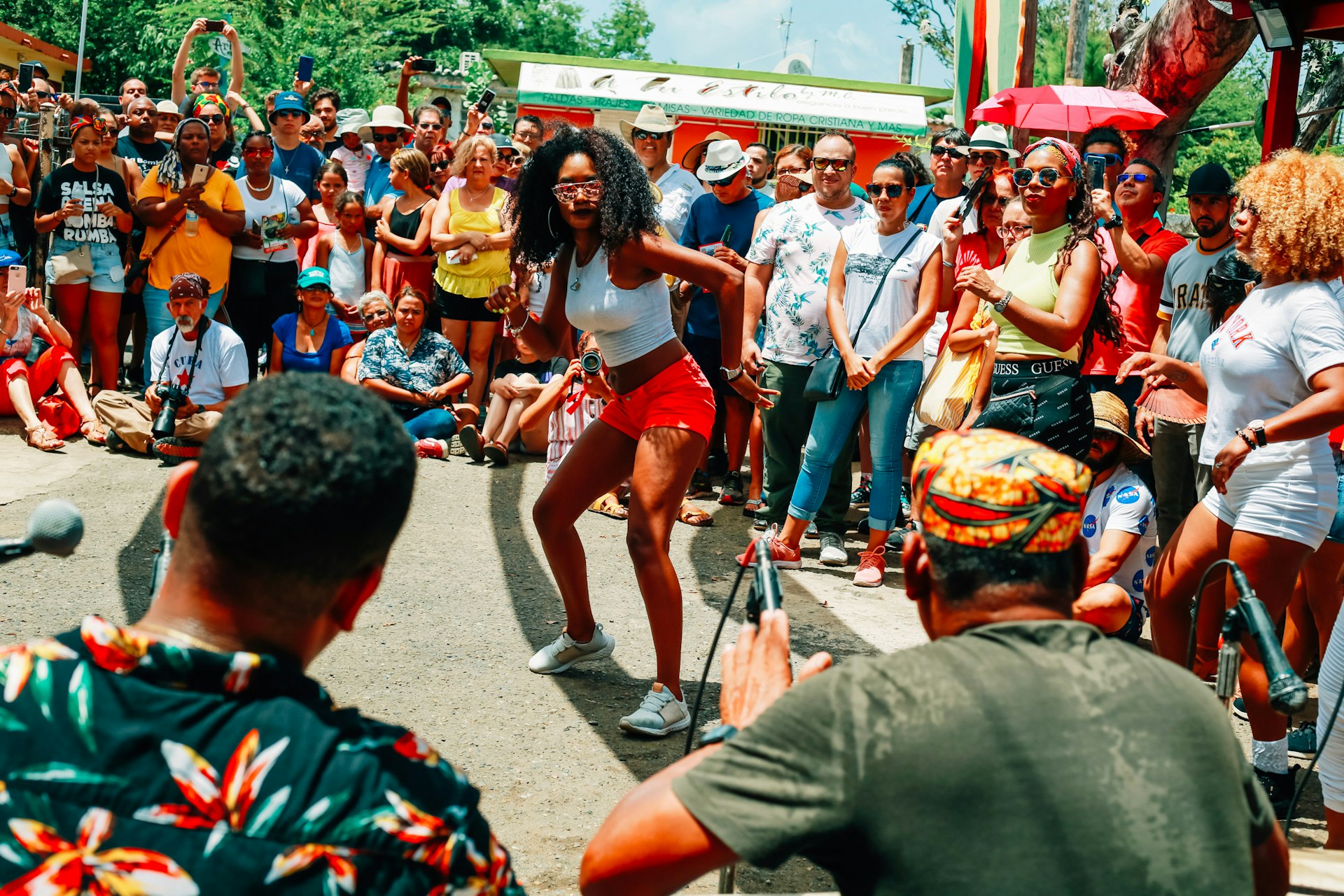 A woman wearing red shorts and a white crop top dances in the center of a circle as a group of musicians play the drums and other folks watch. 