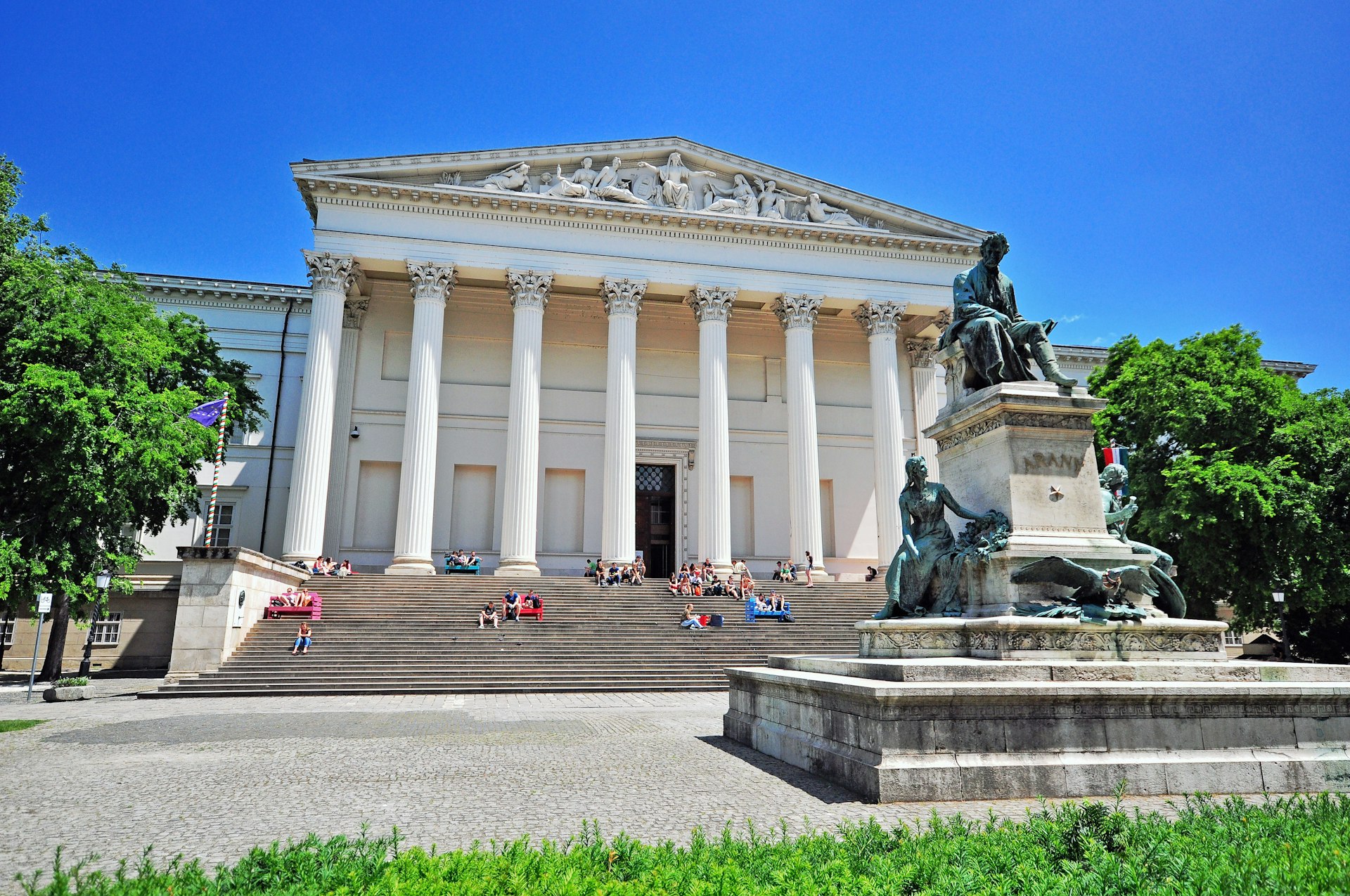 Facade of Hungarian National Museum in Budapest, a large white colonnaded building with people sat on its wide steps
