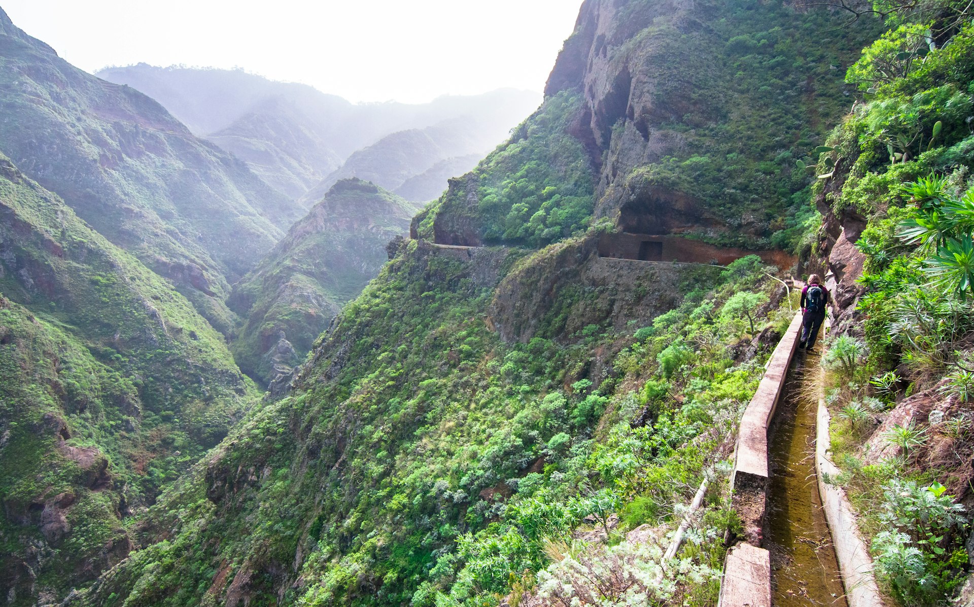 A hiker walks down an old aquaduct that winds along the side of jagged green mountains in Tenerife