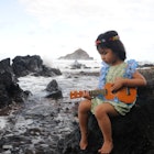A little girl sits on rock next to the ocean, wearing a Hawaiian lei and playing a ukelele. (Maui, Hawaii)