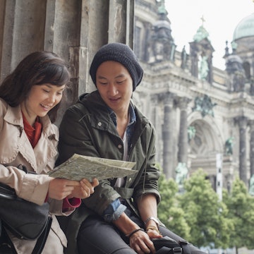 A couple looks at a map while leaning sitting next to stone pillar. In the background is the Berlin Cathedral.