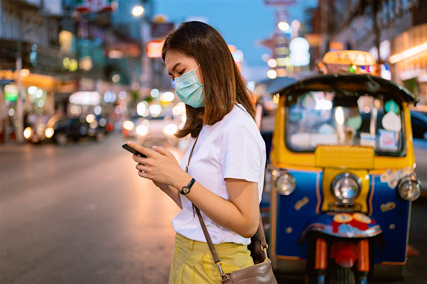 A young woman wearing a mask looks at her phone on a Bangkok street