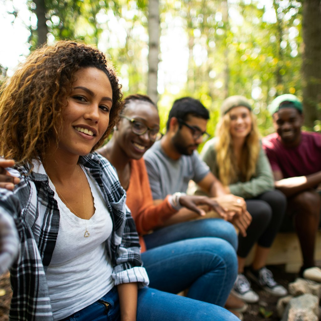 A group of multi-ethnic young adults sit on a wooden bench around a camp fire. They are all dressed casually in fall clothing and sitting around a fire pit.  The mixed race woman on the ed is holding out her camera to take a selfie of the group.