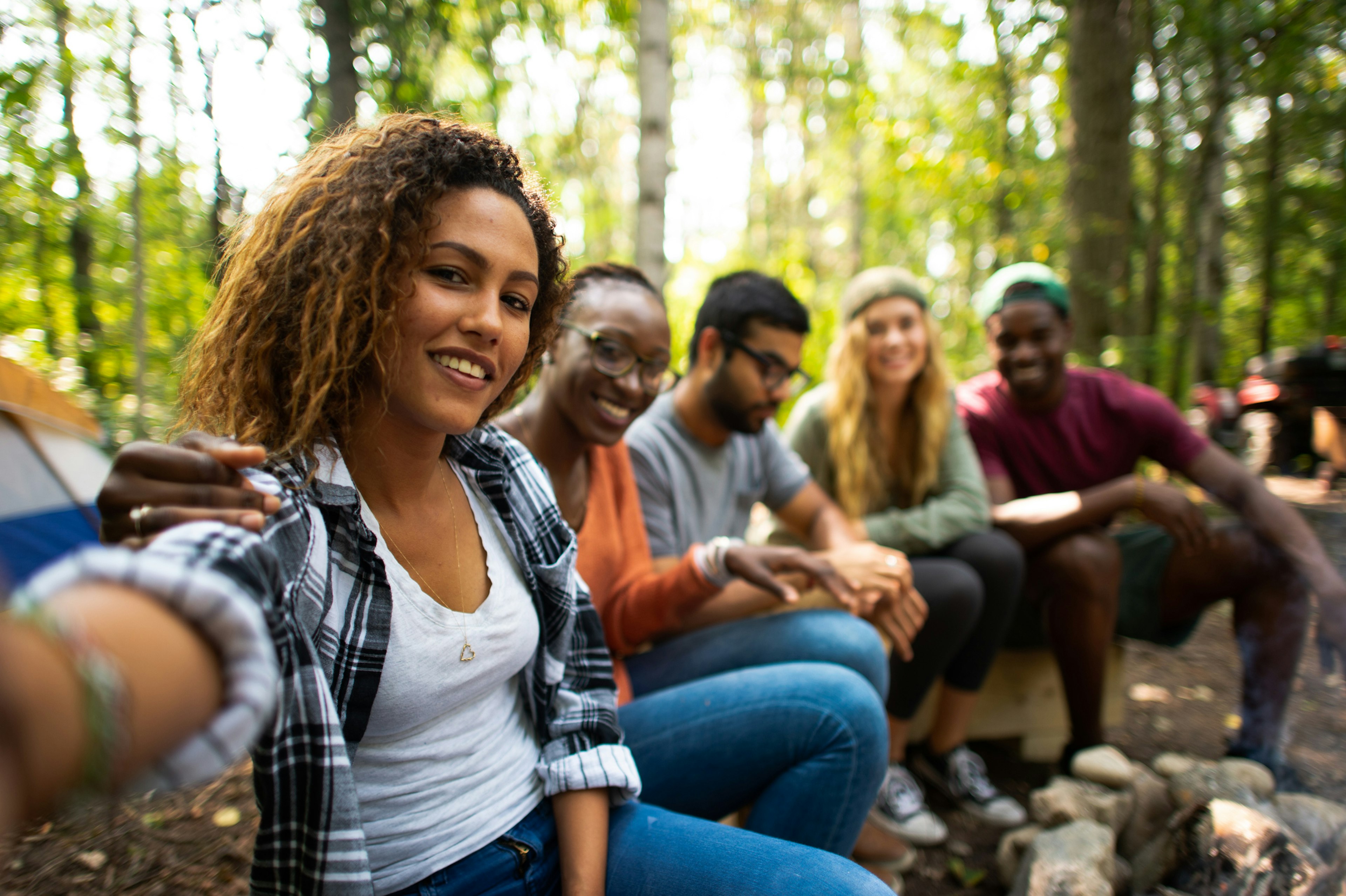 A group of multi-ethnic young adults sit on a wooden bench around a camp fire. They are all dressed casually in fall clothing and sitting around a fire pit.  The mixed race woman on the ed is holding out her camera to take a selfie of the group.