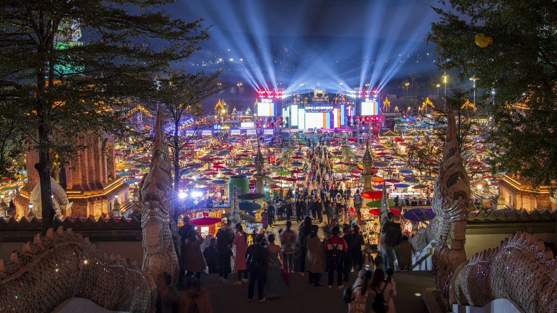 A night shot of a busy market, with a huge colorful light display coming from a nearby building