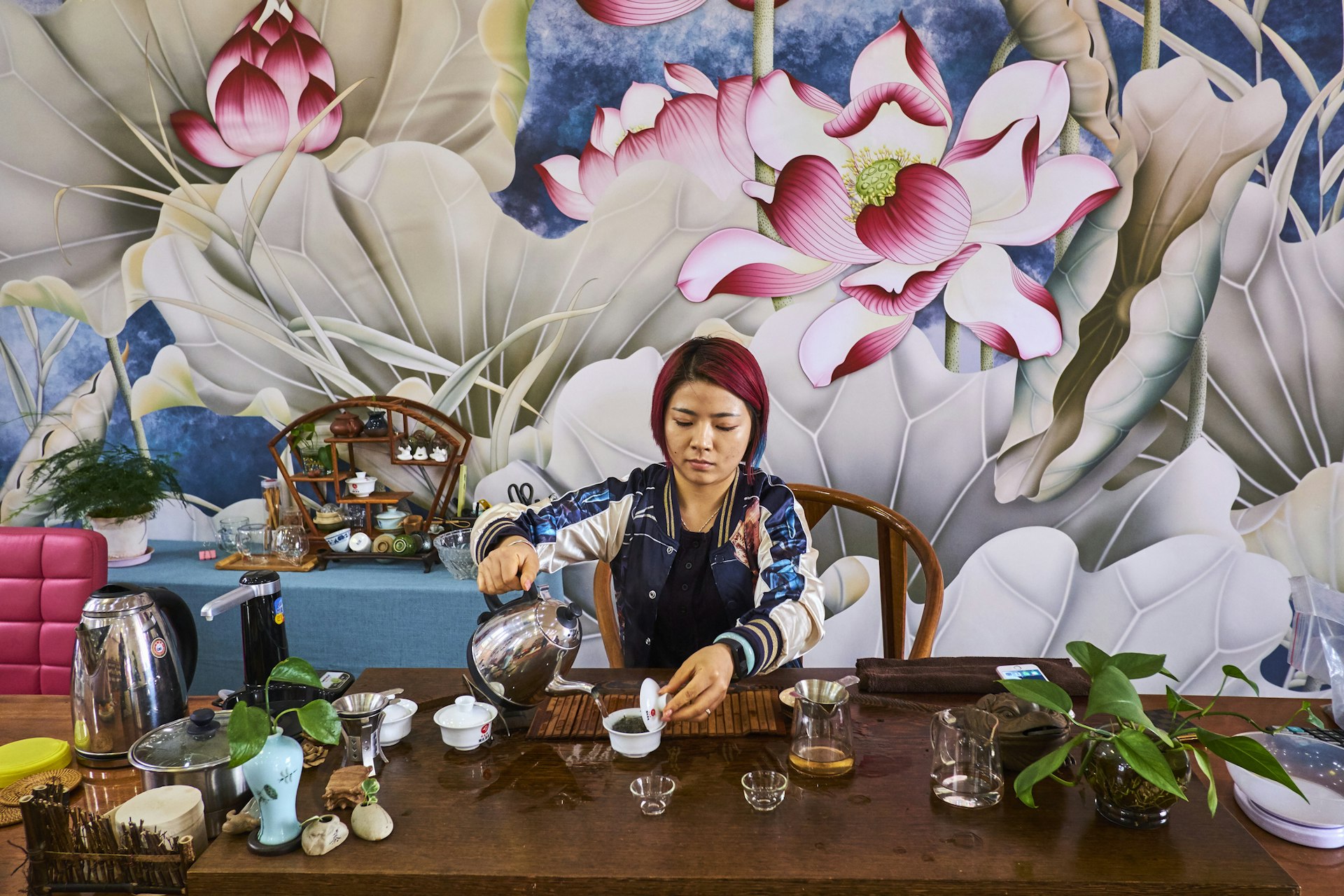 A woman with pink hair pours tea from a teapot in a shop with a large lily painted on the wall behind her