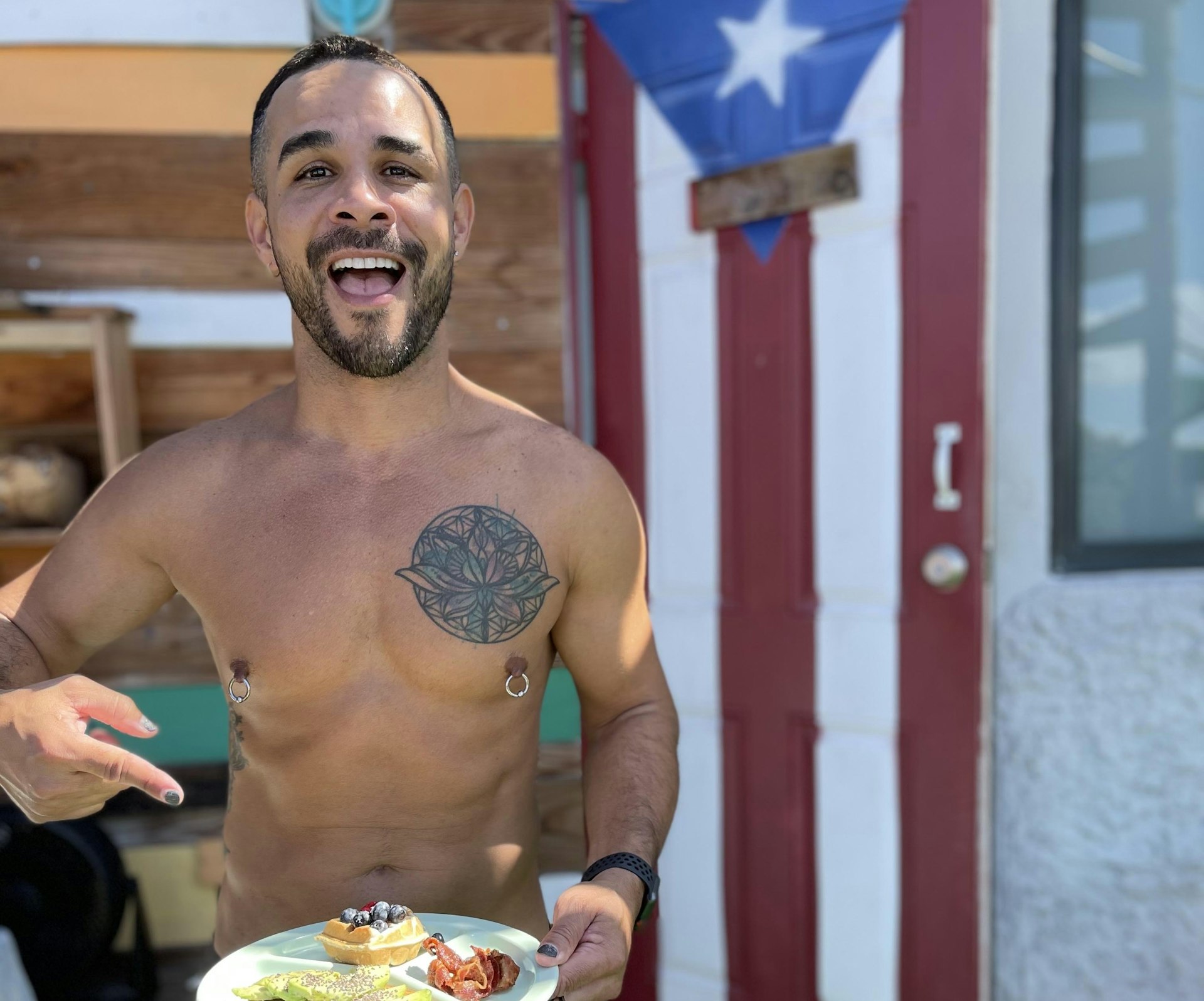 A man points to a plate of food while standing in front of a wooden door painted in the Puerto Rican flag.