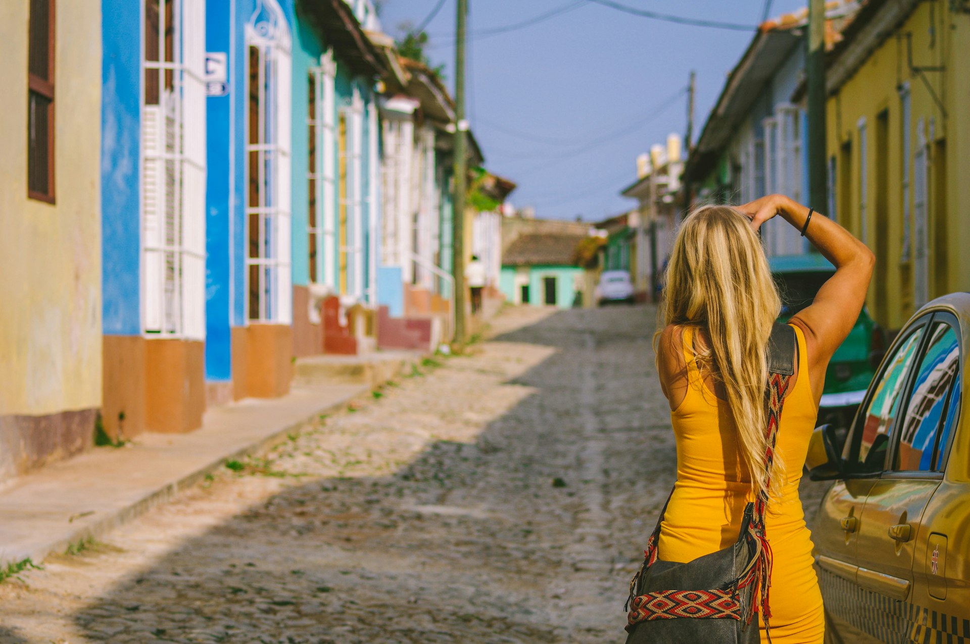 Young blonde girl photographing a street in Trinidad with a colorful yellow dress contrasting with the old empty street popular for tourists and locals