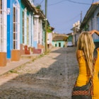 A young blonde woman photographing the streets of Trinidad, Cuba in an early morning light. The street is empty and some of the houses are brightly painted in blue and yellow and other hues.