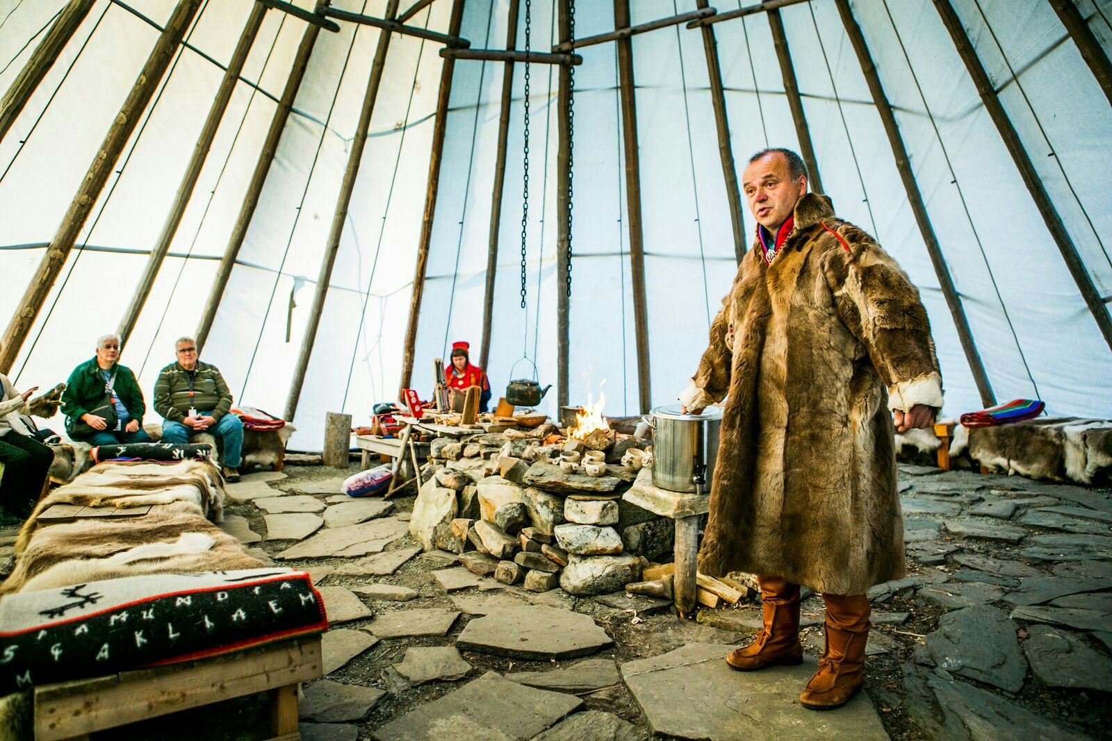 Davvi Siida talks to a group during a Sami Experience tour. He wears a large fur coat and is standing next to an open fire.