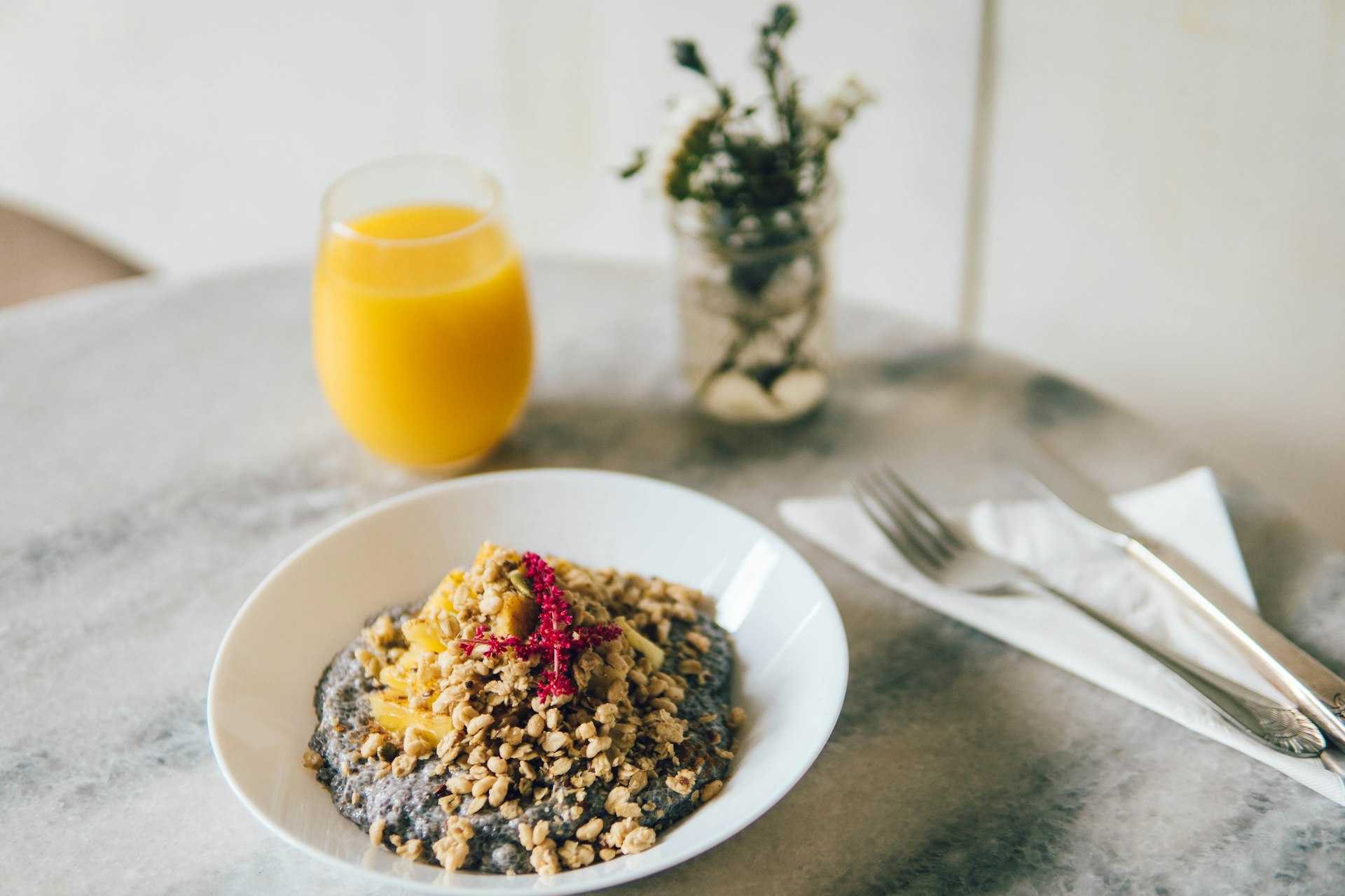 A bowl of beautifully arranged chia pudding with a glass of orange juice and cutlery in the background