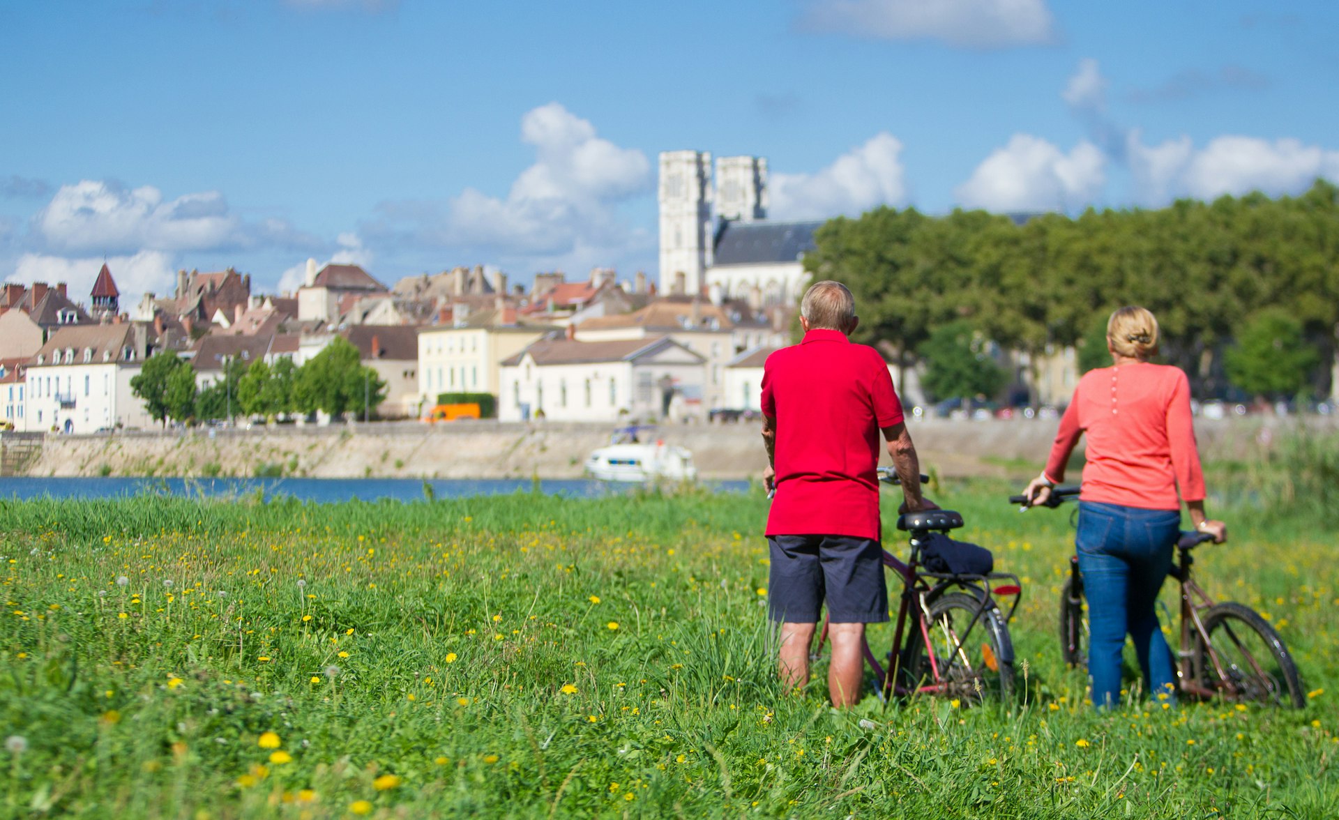 Rear view of an older man and woman with bicycles stood in a grassy field against a blue sky looking across a river at the buildings of Chalon-Sur-Saône in France