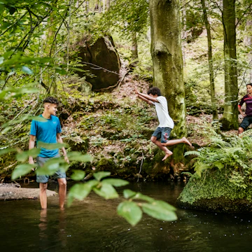 A young boy jumping off a rock into a river while out hiking a woodland trail with his family in the afternoon.