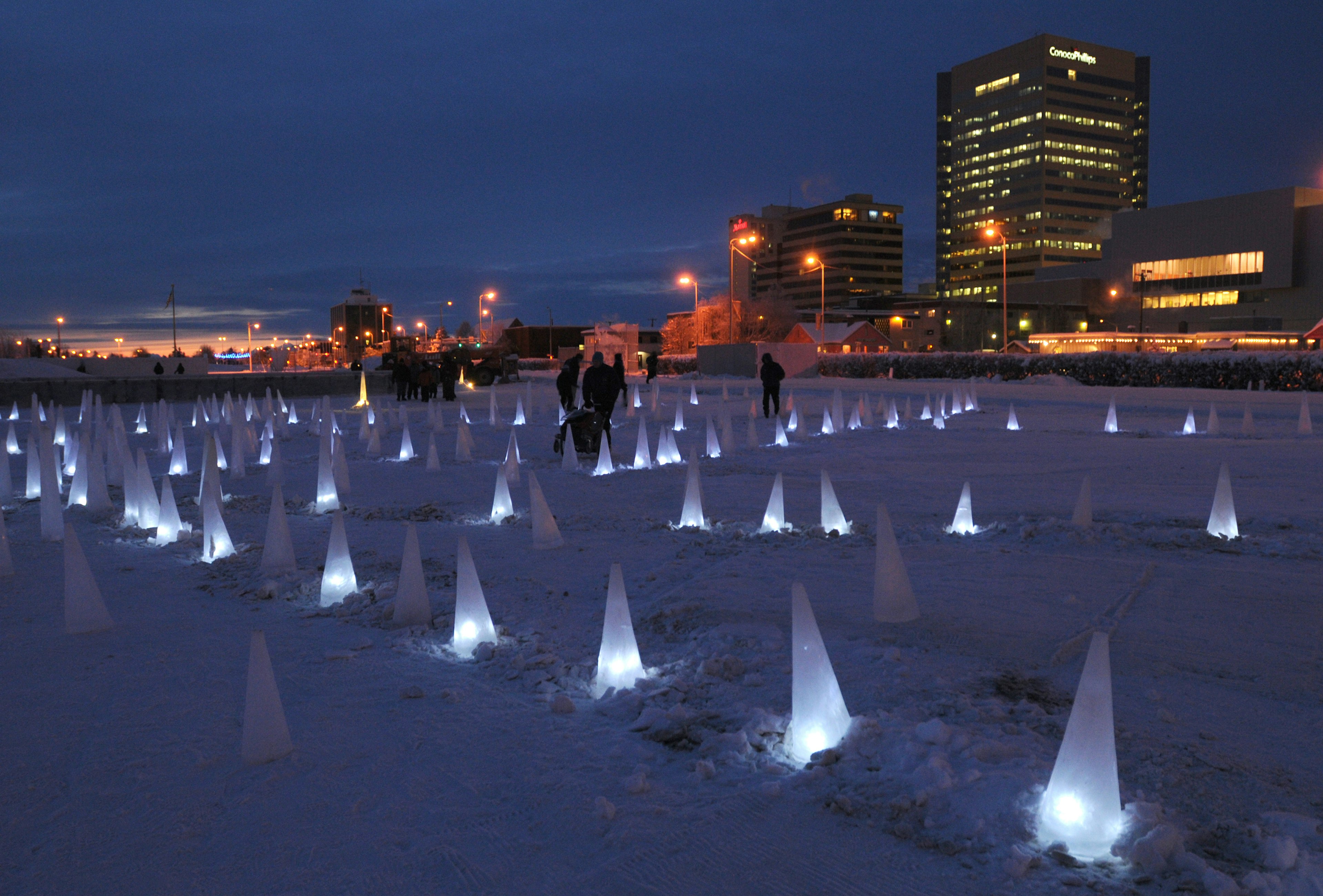 Illuminated ice cones at night on a snowy open area with the Anchorage skyline beyond, part of an art installation called