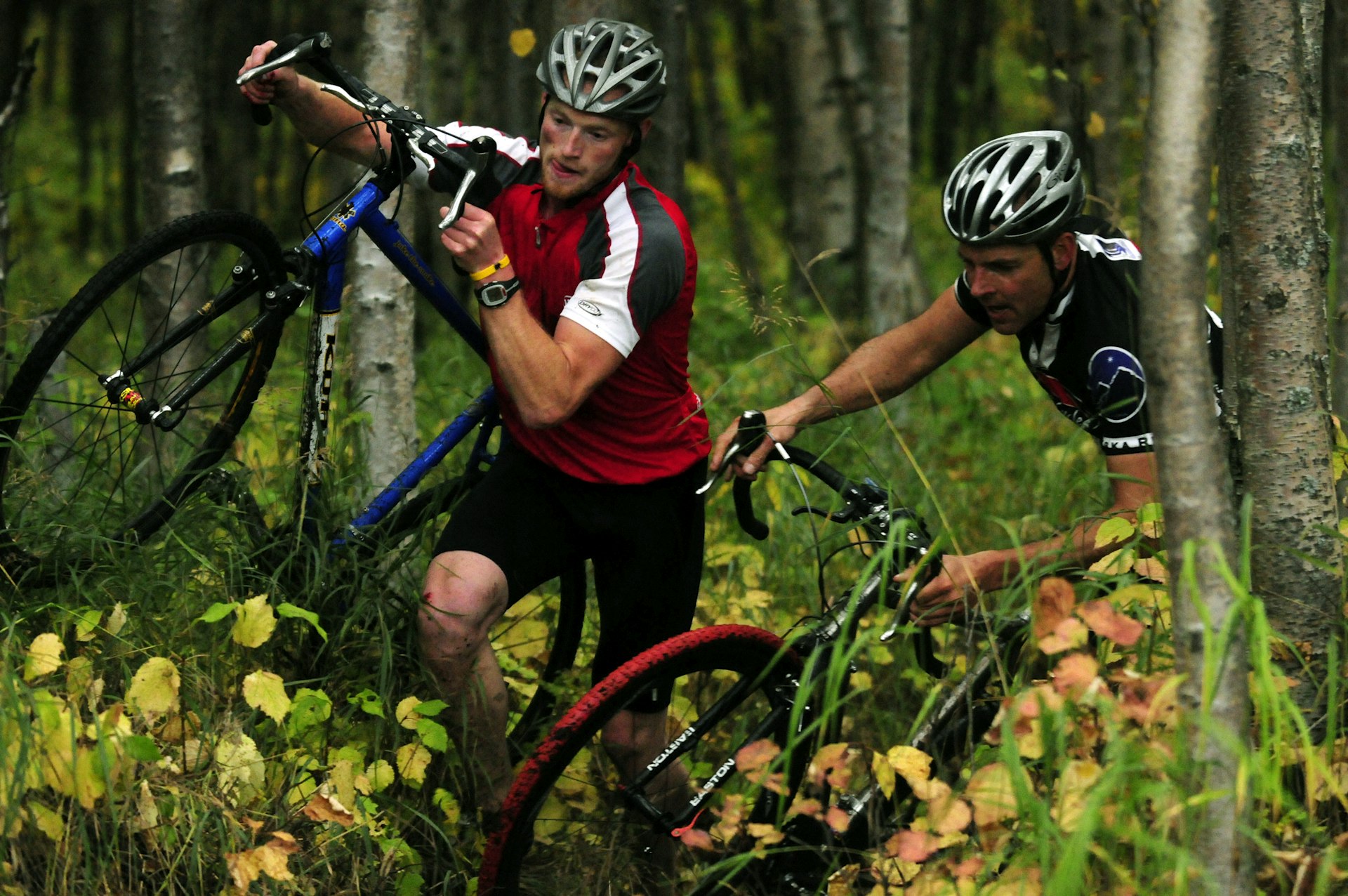 Two cyclists in the woods carry their bikes during a cyclocross race at Russian Jack Springs Park