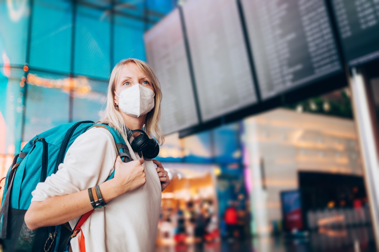 Portrait of a young woman checks the arrivals and departures board at the airport. She wears a face mask for protection during a Coronavirus pandemic..New normal lifestyle for public transport after Covid-19