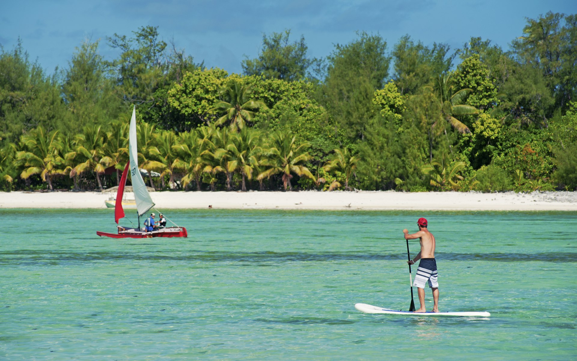 A man stand-up paddle-boarding on the crystal clear water of Muri Lagoon, with a small boat in the background