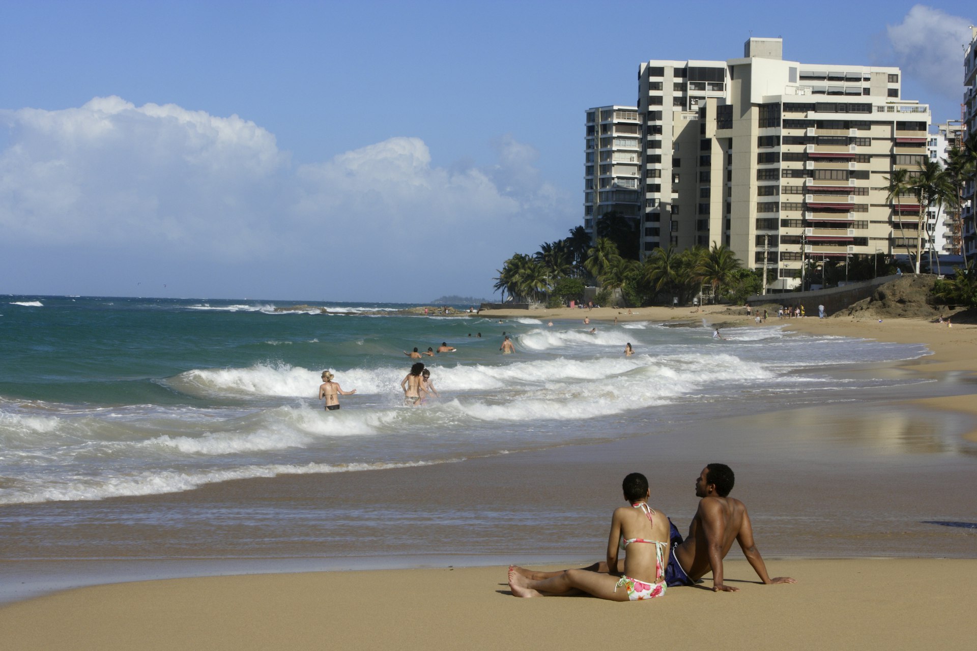 Tourists relaxing on the beach and in the water at Condado, with high-rise buildings and palm trees in the distance