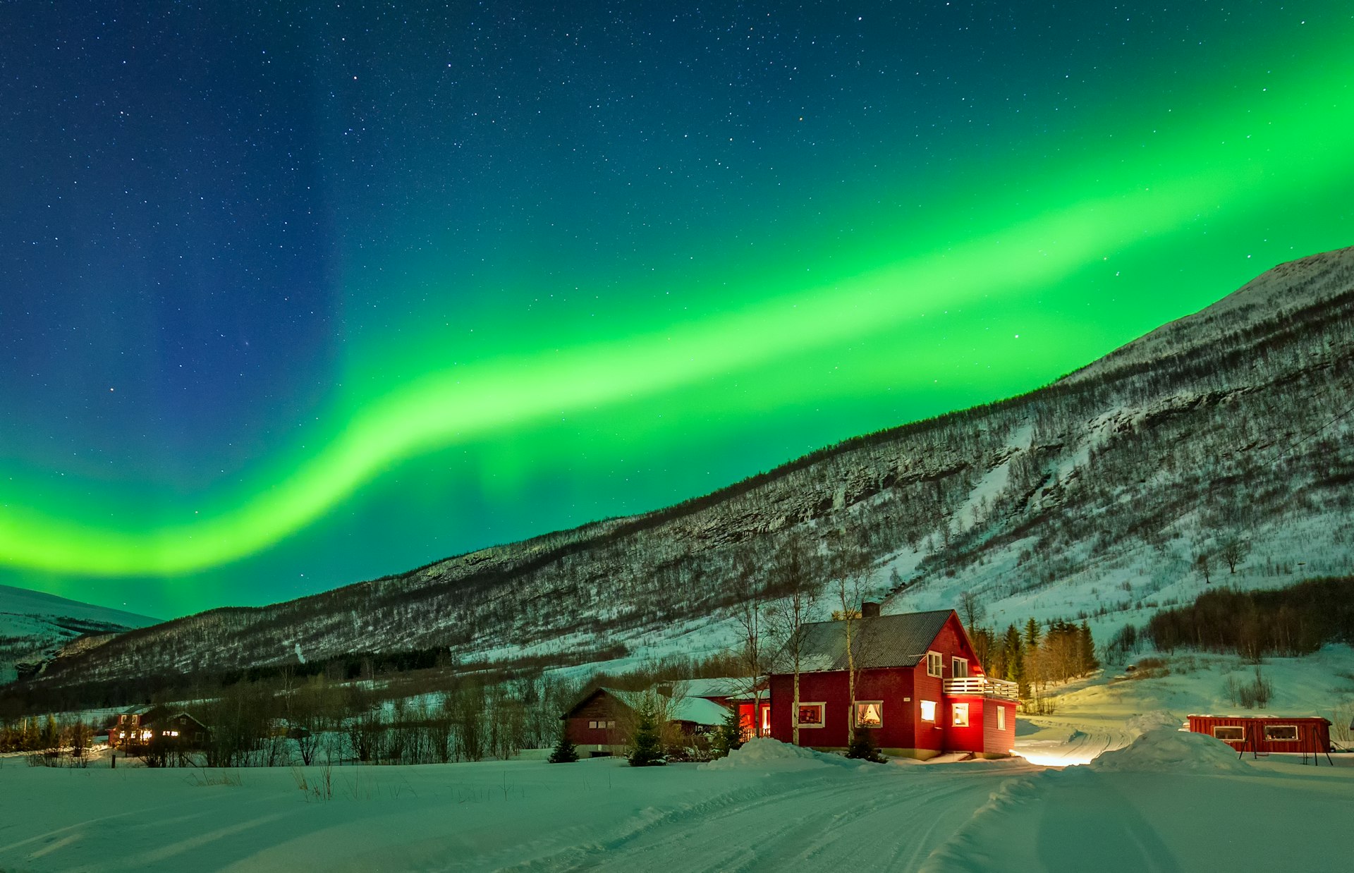 A streak of blue-green lights dance across the sky above an isolated red house in Norway