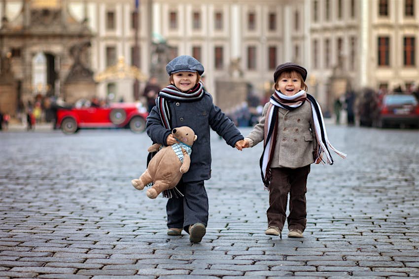 Two boys with hats, scarves and a teddy bear walking the cobblestone streets of Prague