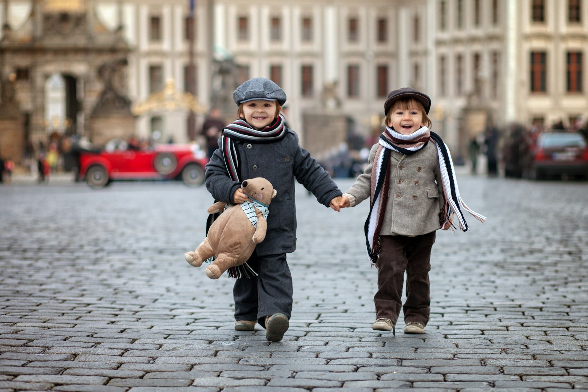 Two boys with hats, scarves and a teddy bear walking on cobblestone streets in Prague