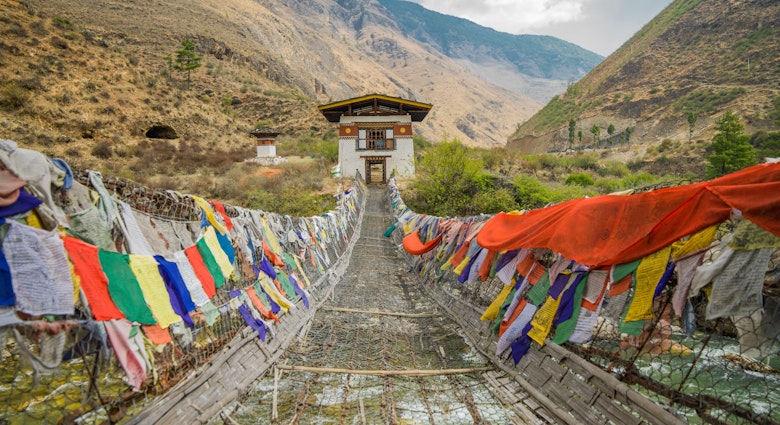 Tachog Lhakhang Iron Chain Bridge with colourful prayer flags on each side of bridge. River crossing. Himalaya mountains in the background.Between Paro and Thimphu Bhutan