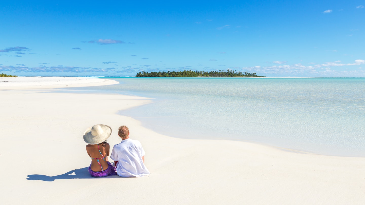 Tourist couple sitting on sandy beach, Honeymoon island, Aitutaki lagoon, Cook Islands, Pacific islands.