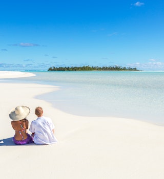 Tourist couple sitting on sandy beach, Honeymoon island, Aitutaki lagoon, Cook Islands, Pacific islands.