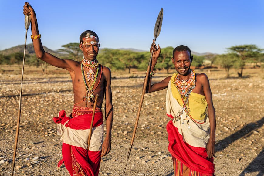 Two Samburu men in traditional wear hold spears and look into the camera