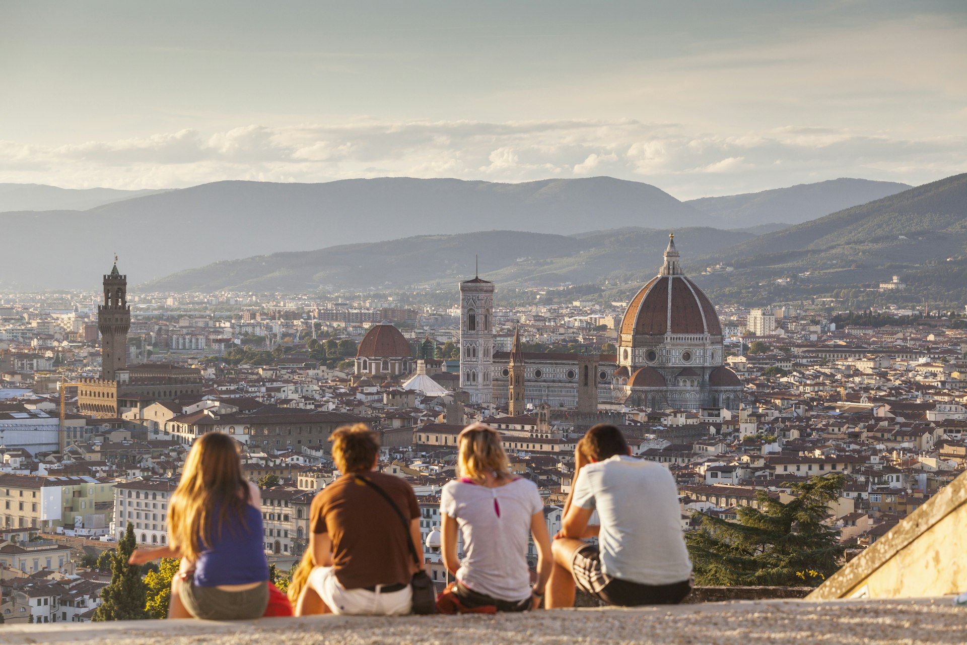 A group of four people sit at a viewpoint looking out over a city. The skyline is dominated by a huge domed church