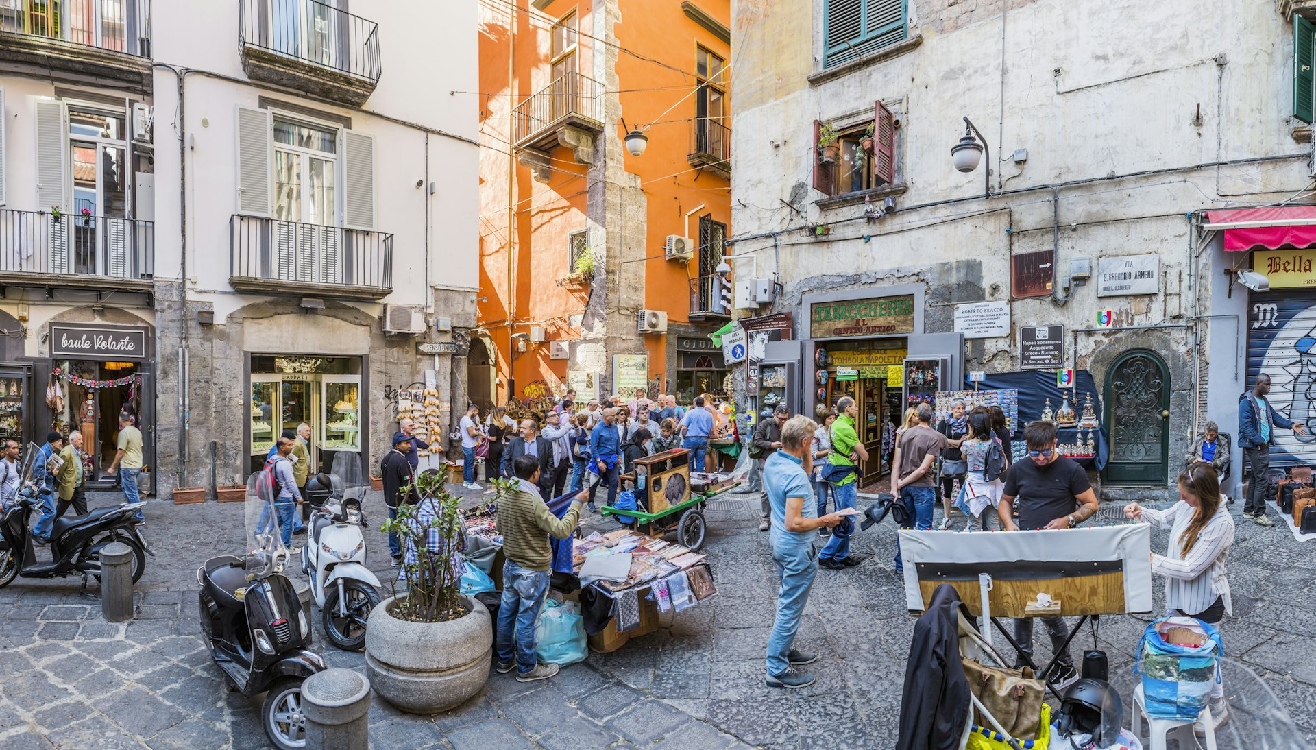 A busy square in an old town of a city, with people milling around and a couple of stalls