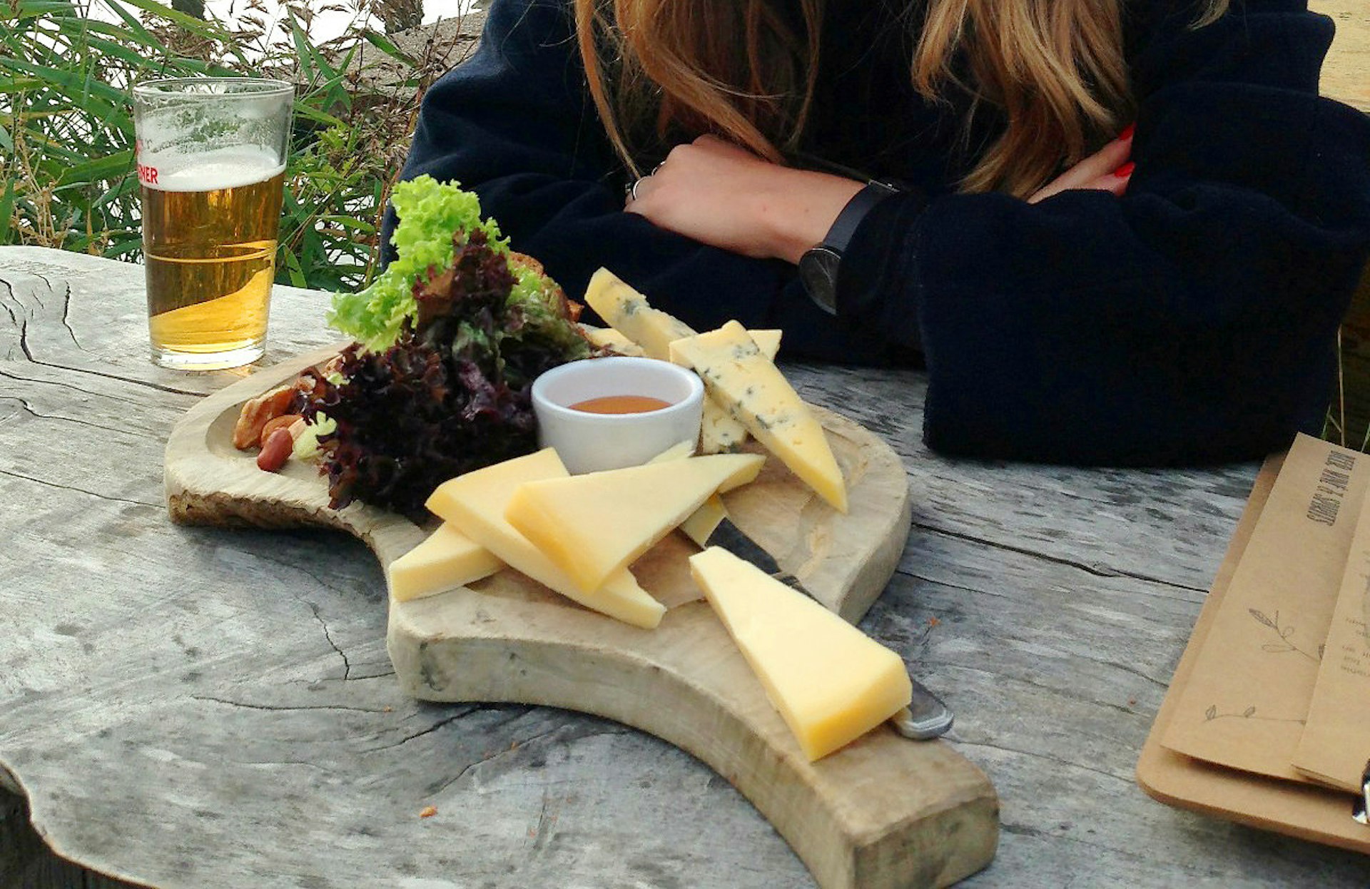A woman with crossed arms sat over a Café de Ceuvel cheese board with a pint of partially-drunk lager on the wooden table
