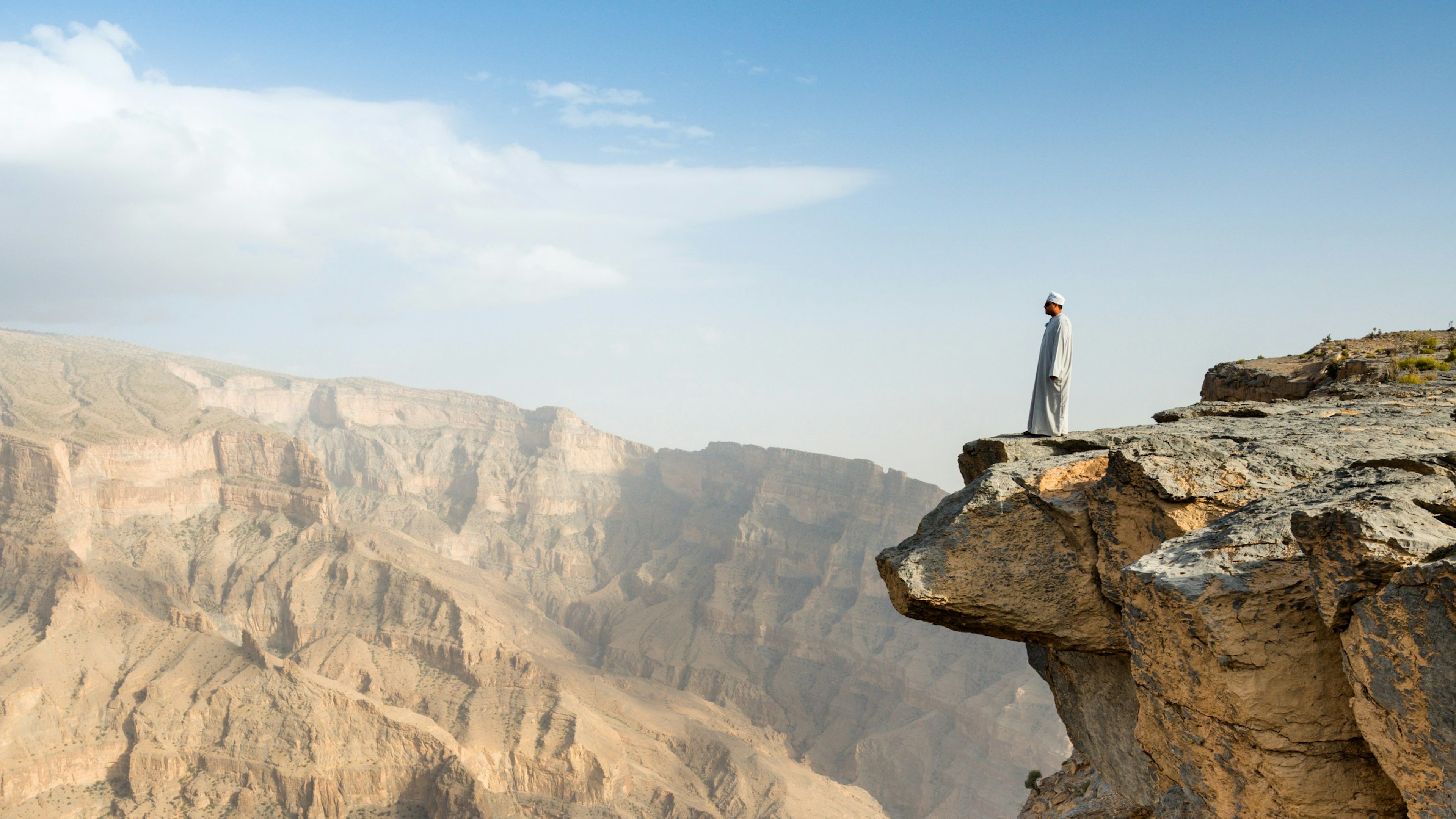 A man in traditional dress stands on the edge of a cliff looking out over Wadi Ghul, Oman, from the top of Jebel Shams mountain