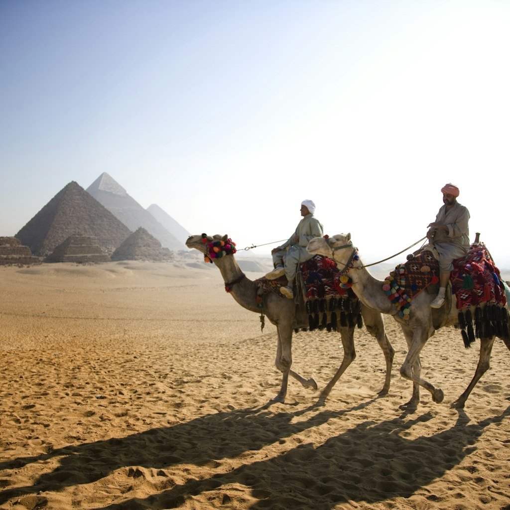 Two men ride on camels in an Egyptian desert. In the background there are a collection of pyramids.
