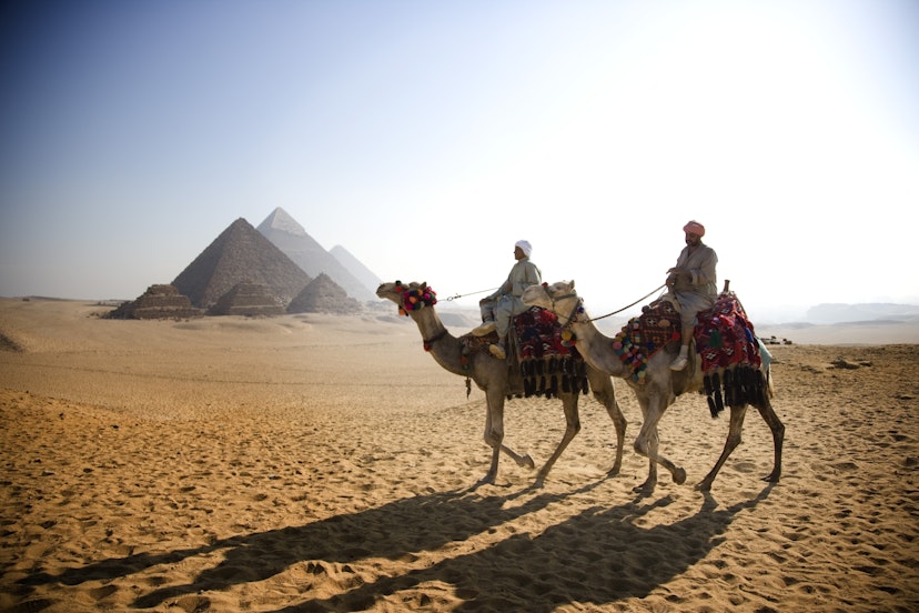 Two men ride on camels in an Egyptian desert. In the background there are a collection of pyramids.
