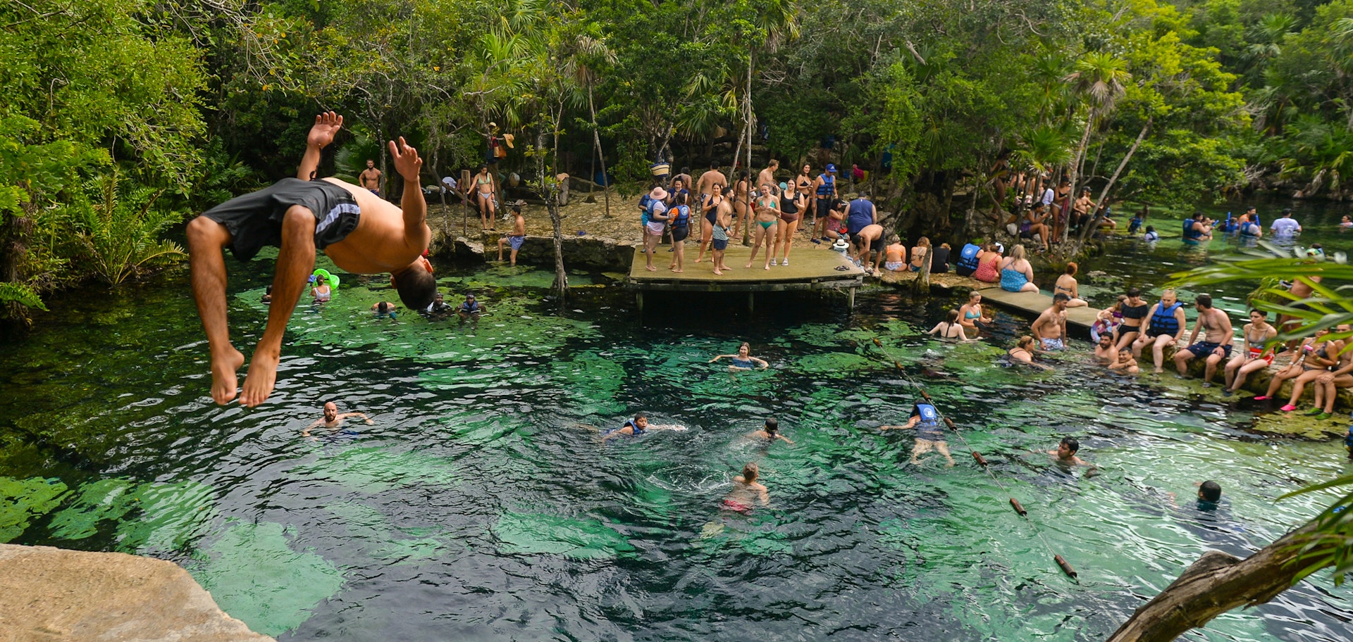 People swim and a boy jumps off a cliff into the water of Cenote Azul, one of the most popular cenotes in the Riviera Maya