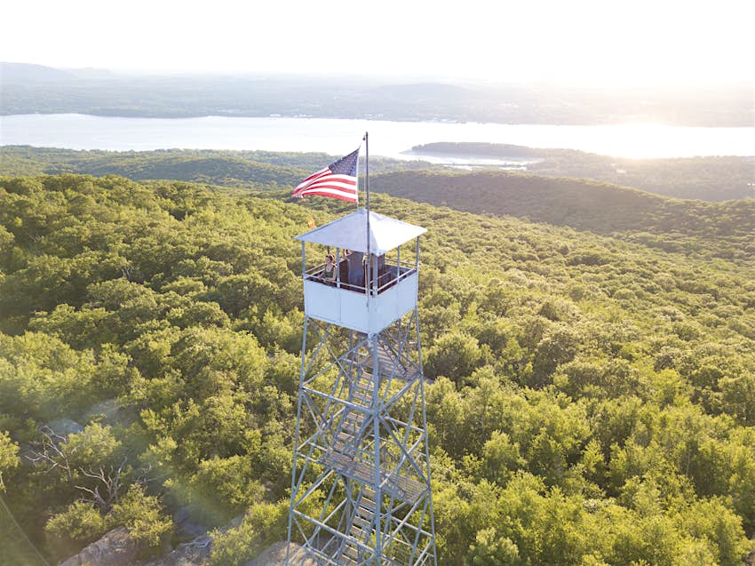 Mt Beacon is one of the best day hikes in the Hudson Valley A woman in a tower with an American flag, looking out over the Hudson River, greenery and mountains