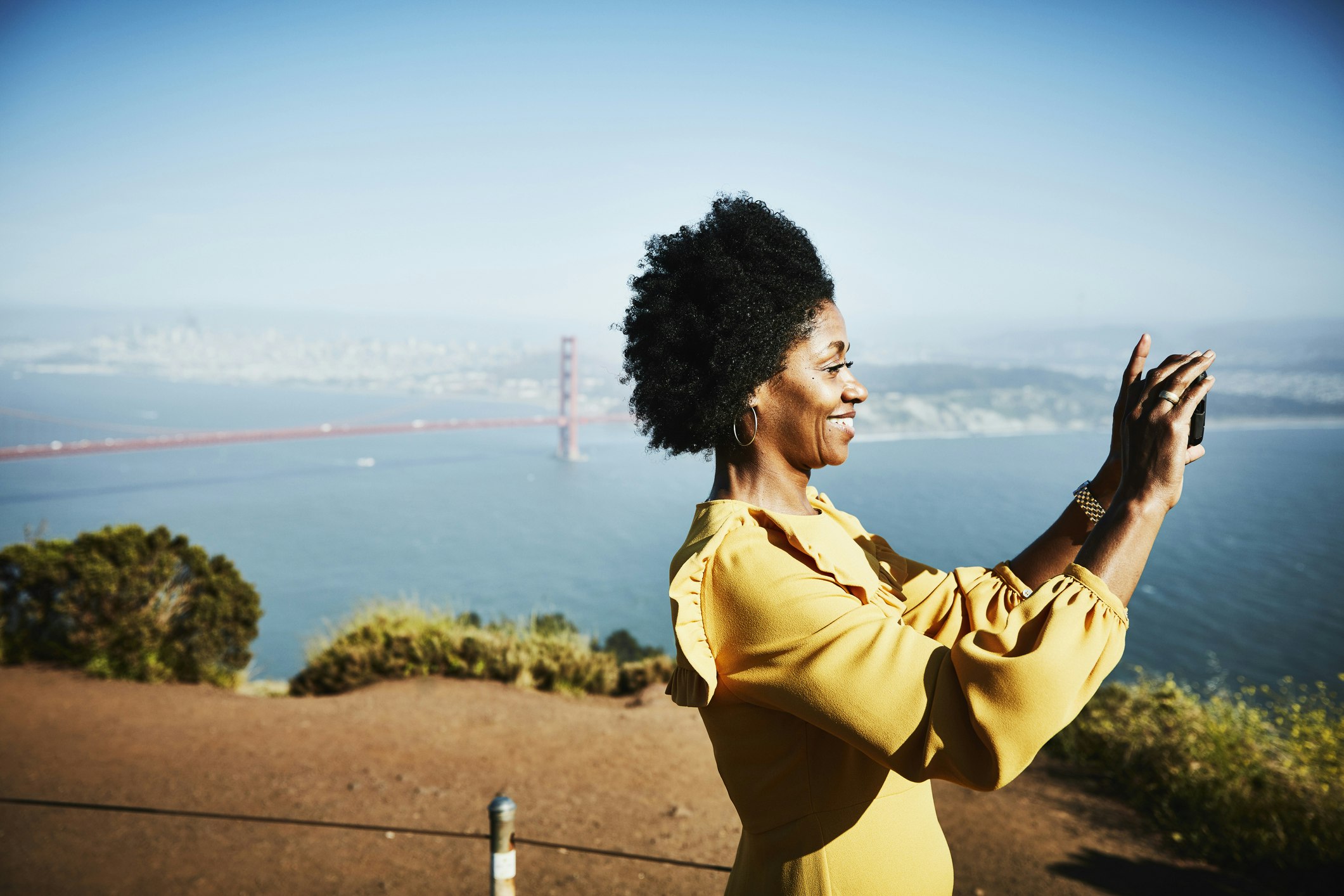 A smiling woman takes a photo with smartphone while standing at a vista at the Golden Gate Bridge above San Francisco, California