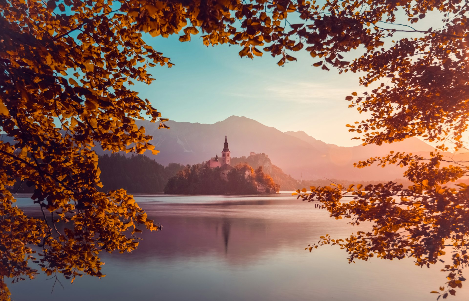 Framed by autumn leaves, Bled Island juts out of Lake Bled at sunrise with the Church of the Assumption with Castle