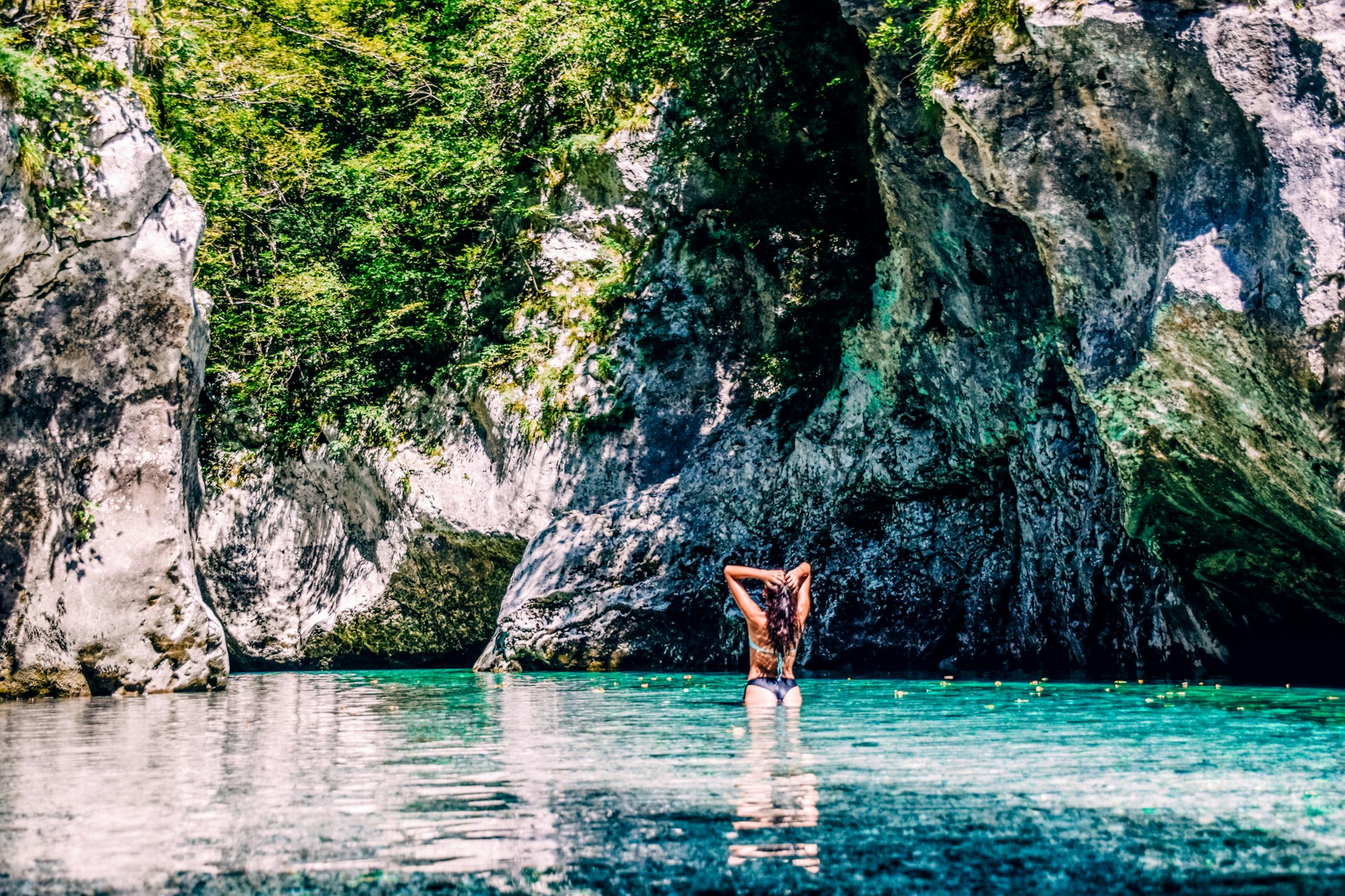 A woman slicks back her wet hair after swimming in the turquoise Soča River with hulking rocks in front of her