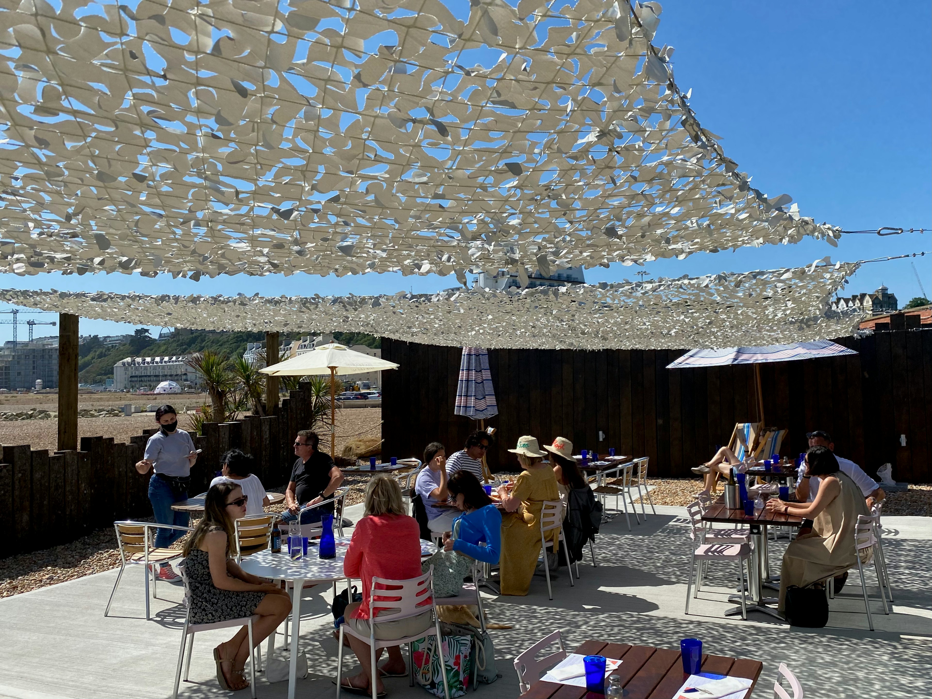 Tables full of people, sitting under a shade on a patio on a bright sunny day