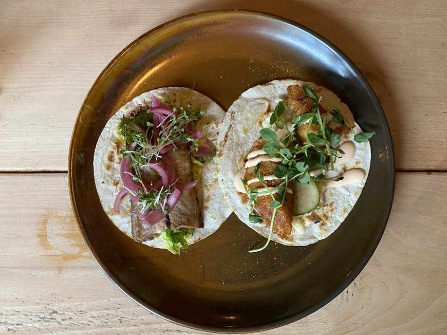 Två tacos på en stor tallrik, en med bringa, syltad lök och groddar, den andra med stekt fisk, kryddig majonnäs, slaw och groddar