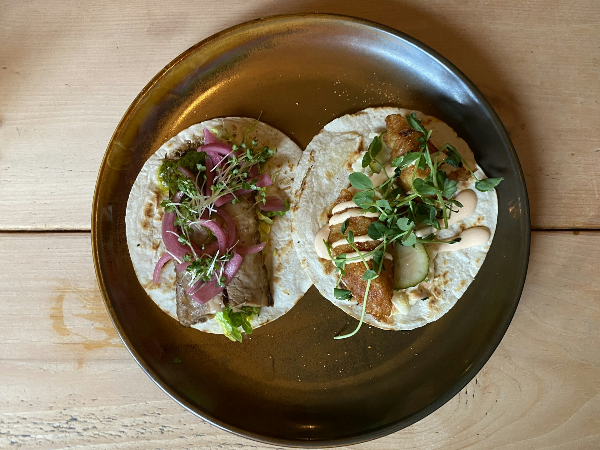 Two tacos on a big plate, one with brisket, pickled onions and sprouts, the other with fried fish, spicy mayo, slaw and sprouts