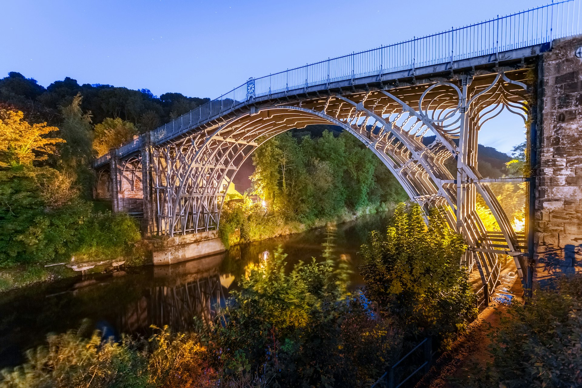 A huge iron bridge spans a gorge; it's shot at dusk with a blue hue to the surrounding light