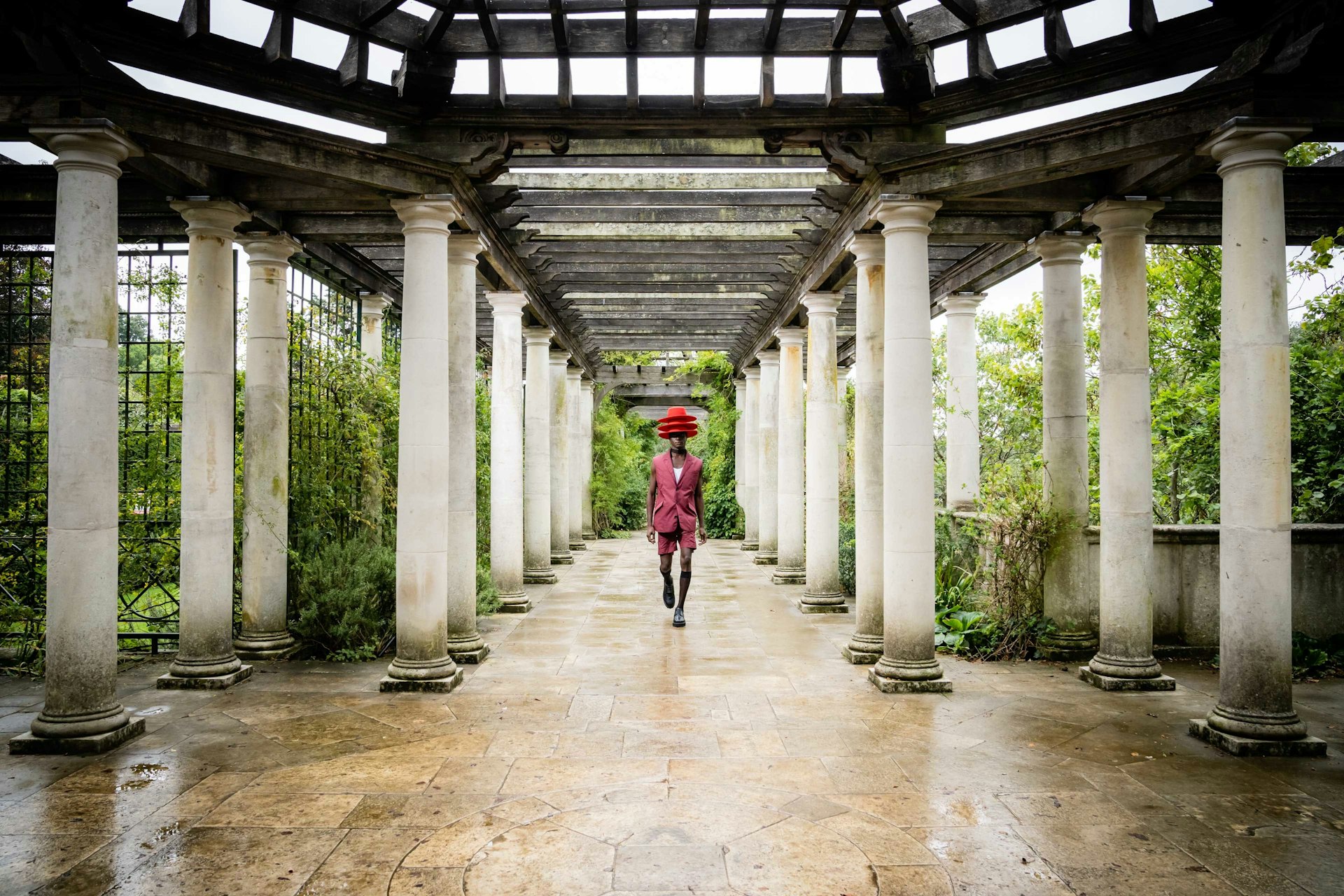 A black male model wearing three red hats strides down a walkway lined with columns