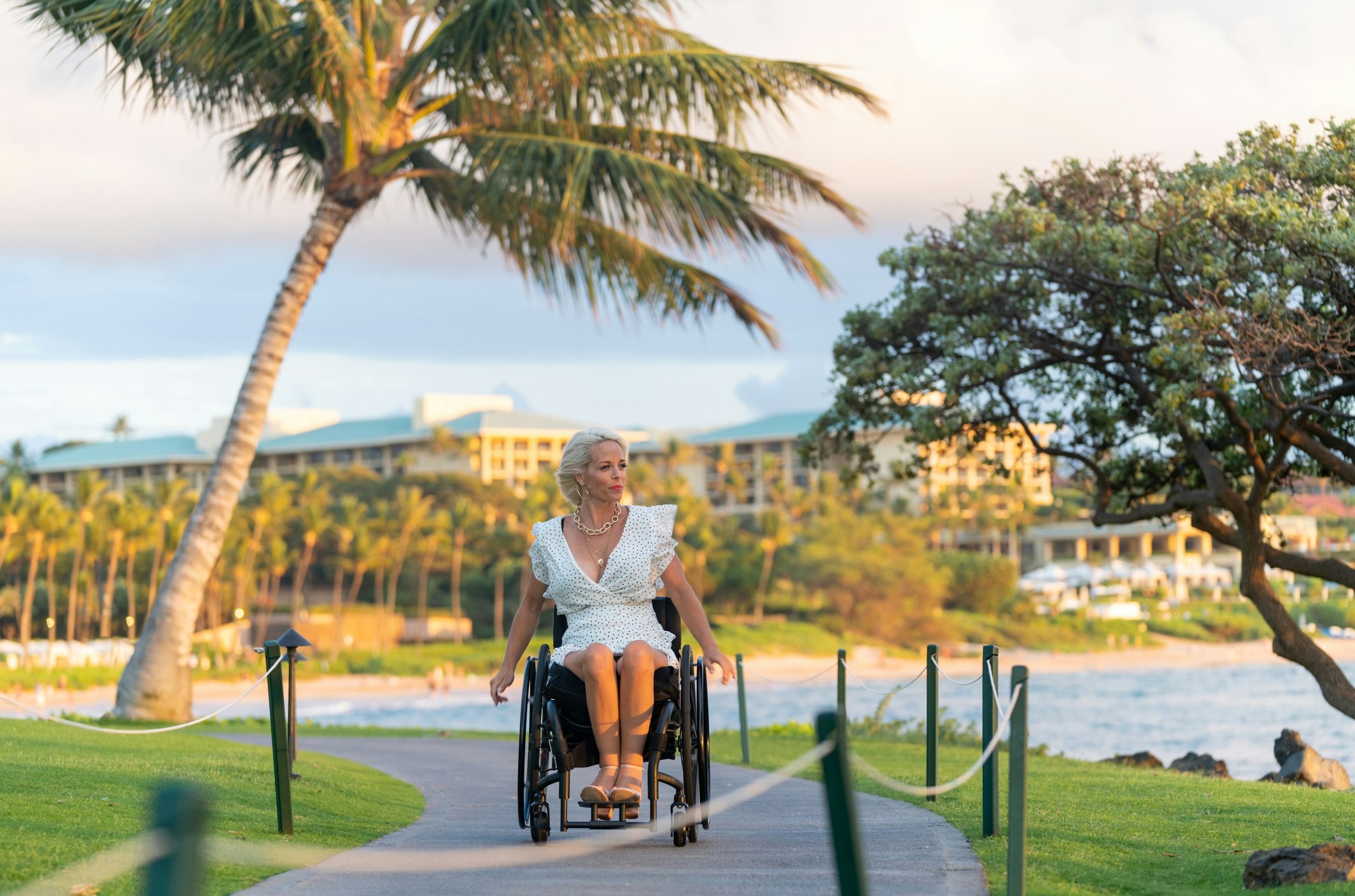 A white woman in a white dress wheels her wheelchair along a boardwalk in a tropical destination