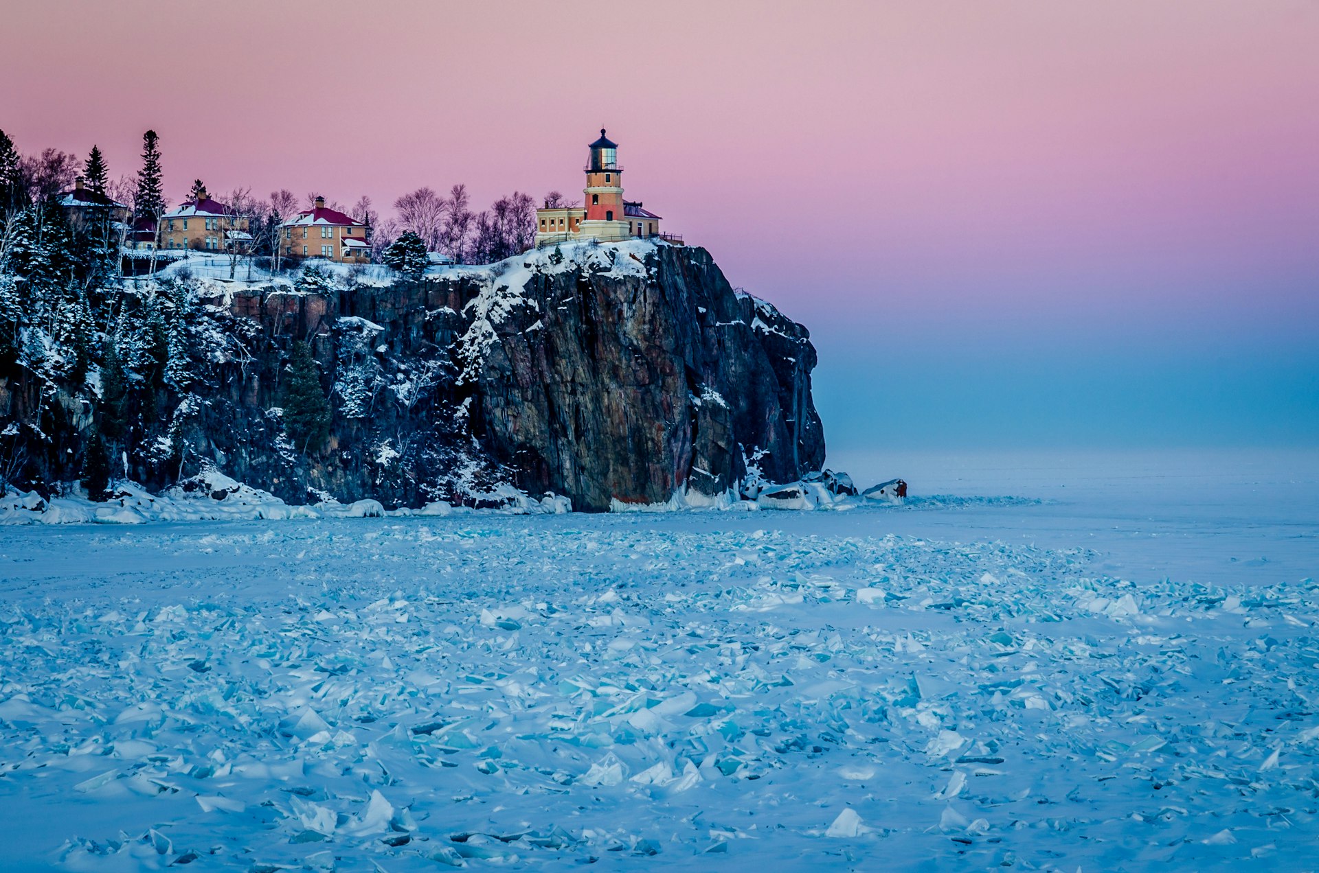 A lighthouse on the top of a rocky cliff glows pink the low sunlight. The lake below is frosty