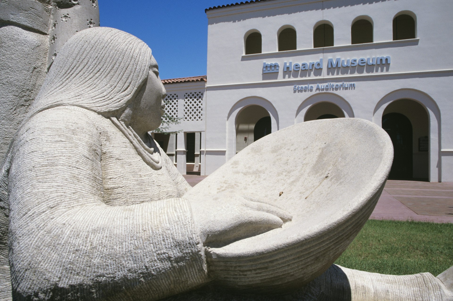 A sculpture of a Native American woman with a bowl sits outside the white-walled Heard Museum in Phoenix, Arizona, USA