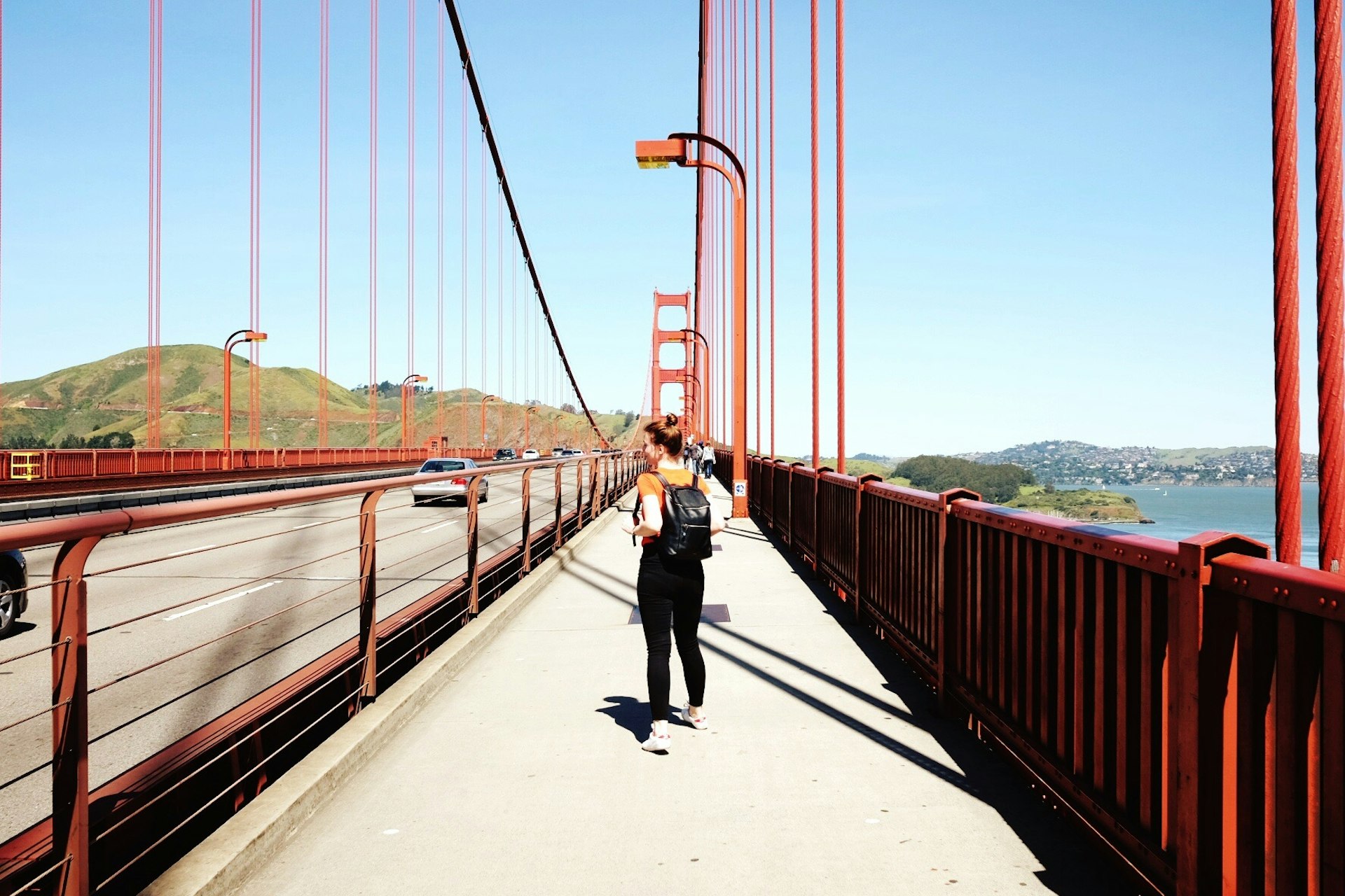A woman walks along a pedestrian path at the side of a road on a large orange-colored suspension bridge
