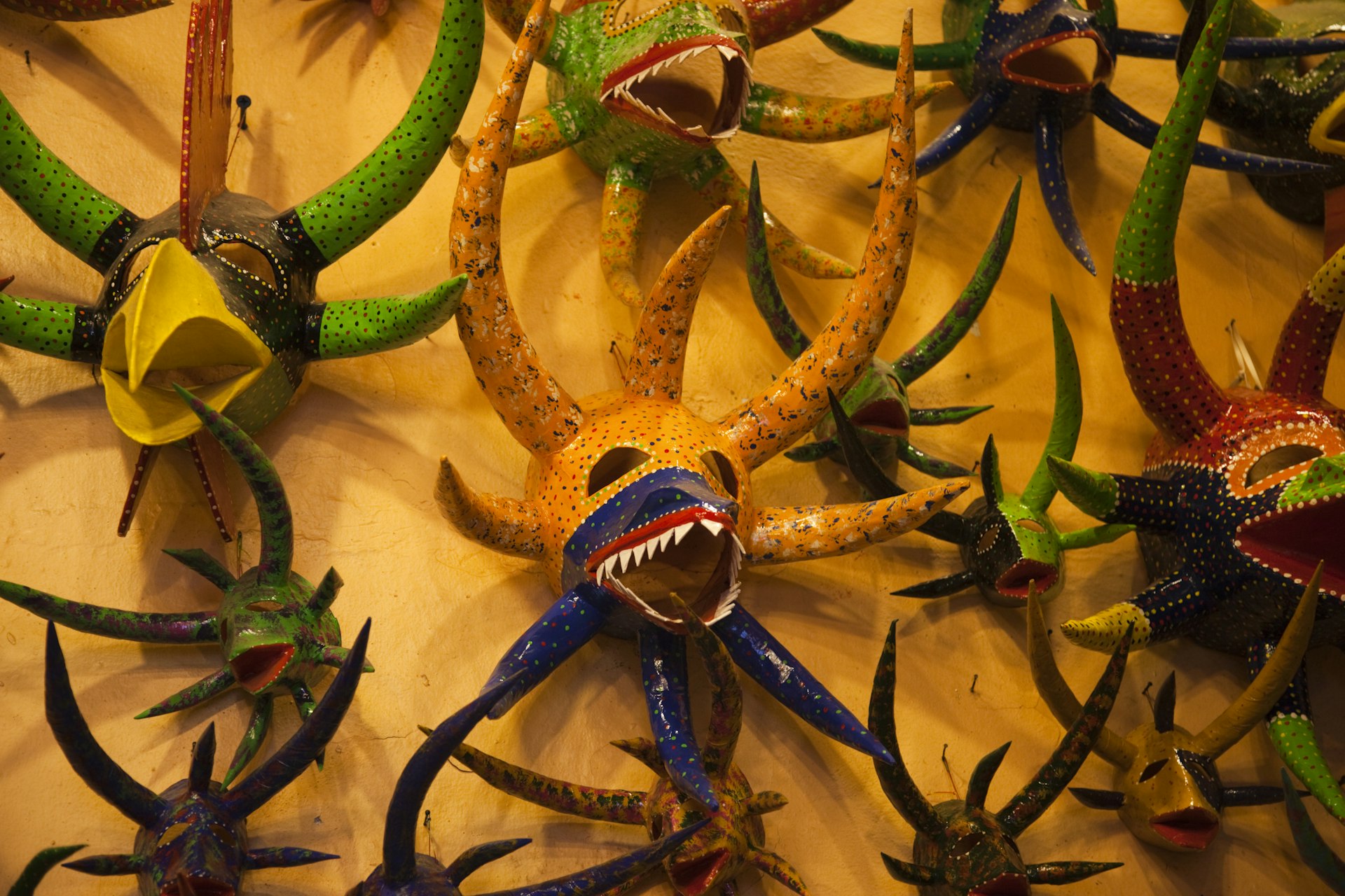 A collection of colorful masks with large horns, some with sharp teeth and another with a large beak are hung on a yellow wall.